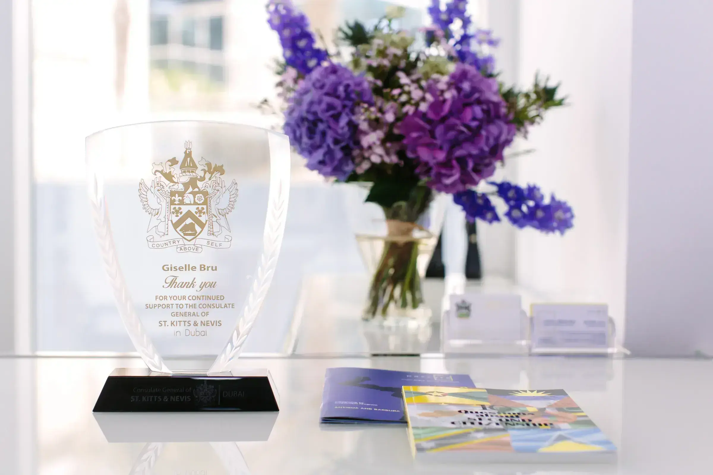 The Consulate General of St. Kitts and Nevis (Dubai) engraved-glass award presented to Giselle Bru, alongside PassPro programme brochures and delphinium florals.