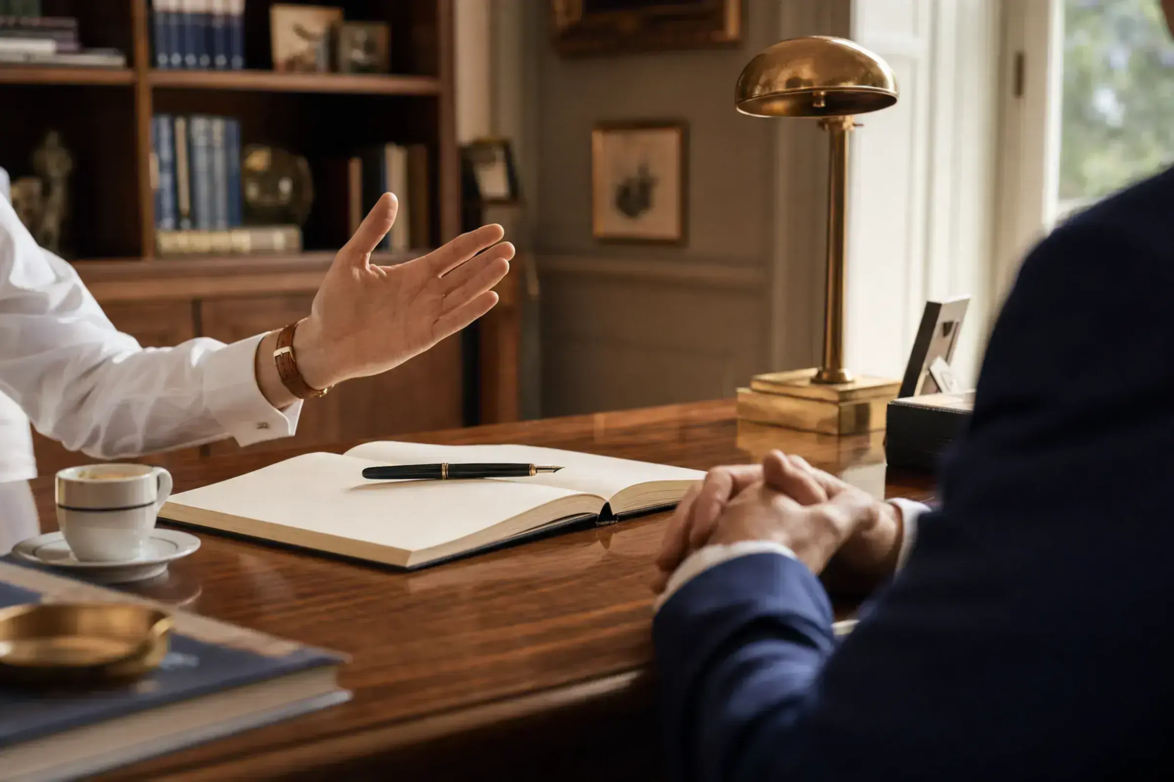 Two advisors sitting across a desk, an open book between them, one with palm extended in explanation — the posture of considered counsel.